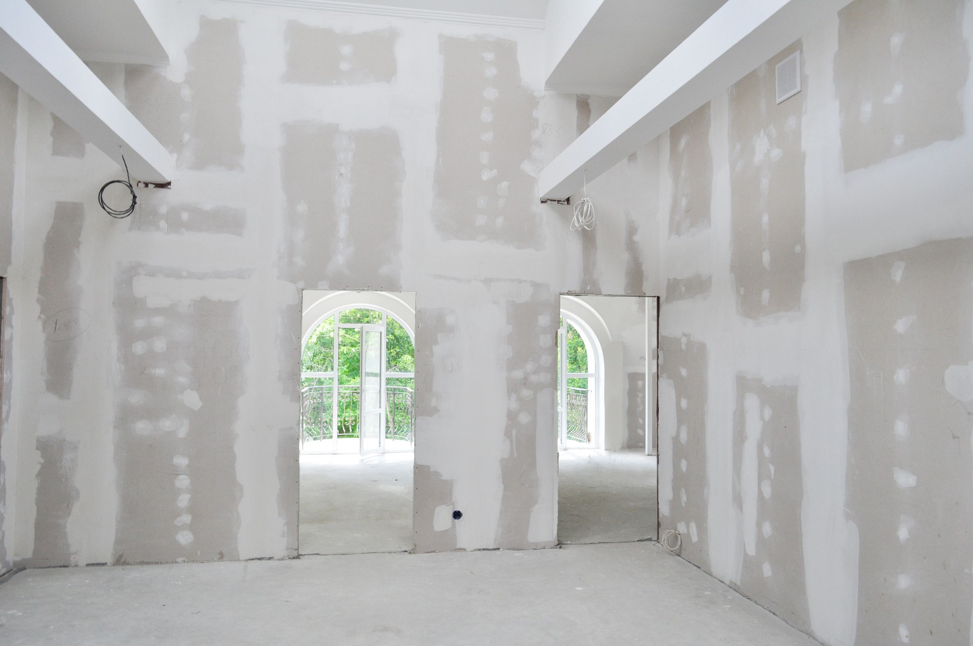 Interior view of a residential building under construction, showing newly installed drywall with taped and mudded seams. The room features high ceilings with exposed beams and arched doorways leading to bright windows that open to a balcony. The smooth drywall finish and clean layout highlight the precision and quality of the interior finishing work in progress. “New drywall installation and finishing in a residential construction project — showcasing smooth seams, high ceilings, and precise interior craftsmanship by a professional construction company.” drywall installation, interior finishing, residential construction, drywall taping and mudding, home renovation, professional construction company, drywall contractor, new home build, interior framing and drywall