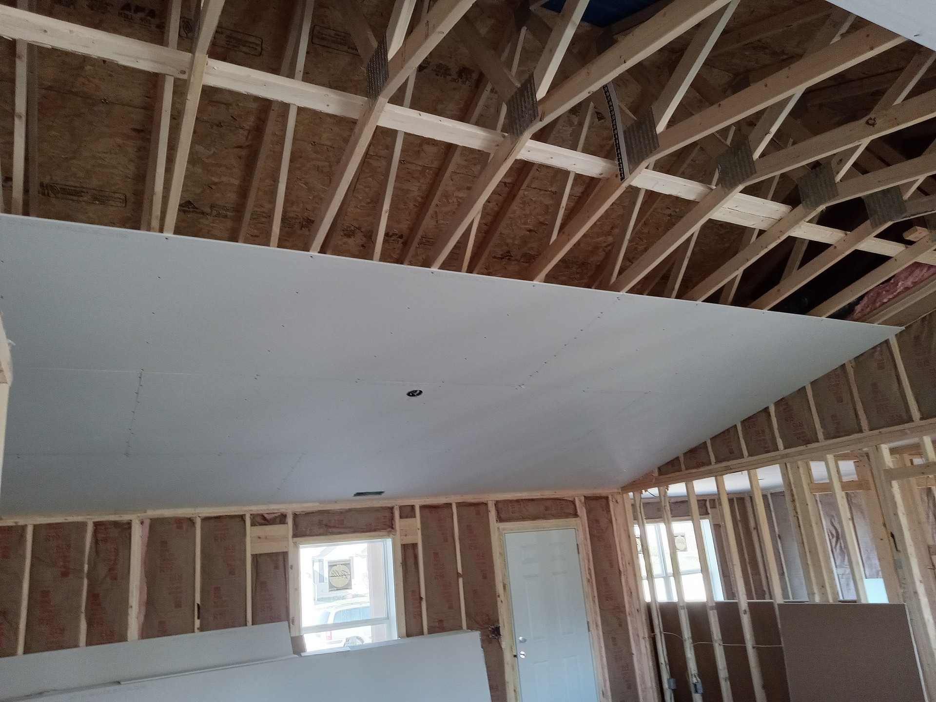 Interior of a residential construction project featuring drywall installation on a vaulted ceiling. The exposed wood framing, roof trusses, and wall insulation highlight the structural phase of the build. A door and window allow natural light to illuminate the workspace. The image demonstrates professional craftsmanship, attention to detail, and quality construction practices during the home-building process. “Residential construction in progress — expert drywall installation and precision framing by a professional construction company, showcasing quality workmanship and structural integrity.” residential construction, drywall installation, framing and insulation, vaulted ceiling build, professional construction company, home building services, quality craftsmanship, new home interior